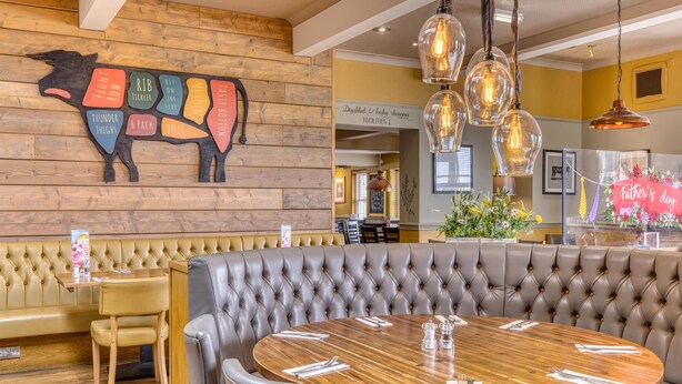 A restaurant interior with a wooden wall, a tufted booth, a round wooden table, and hanging pendant lights. - Premier Inn Arundel (Arundel)