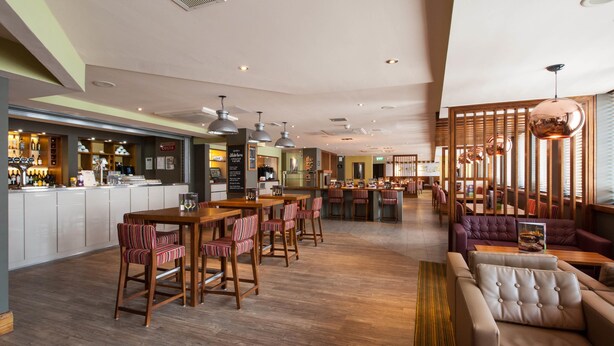 Un grand espace bar avec des tables en bois et des chaises à rayures, un coin salon avec des canapés et un comptoir, et des toilettes visibles au fond.