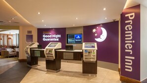 Dans le hall d'accueil du Premier Inn, on trouve un espace d'enregistrement avec un kiosque numérique, un comptoir de réception et un mur orné du logo de l'hôtel et d'une affiche indiquant la « Garantie d'une bonne nuit ».