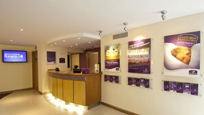 Una zona de recepci ón de hotel con un mostrador de madera, un televisor mostrando 'Welcome to Premier Inn Burgess Hill' y varios carteles promocionales en la pared.