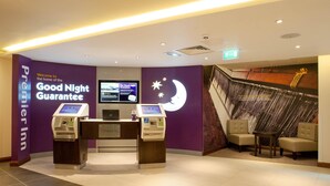 A hotel reception area with a check-in counter, a digital display, and a seating area with two chairs. - Premier Inn Trowbridge (Trowbridge)