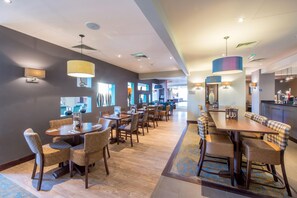 Un comedor de hotel con varias mesas y sillas, una cocina al fondo y una alfombra estampada.