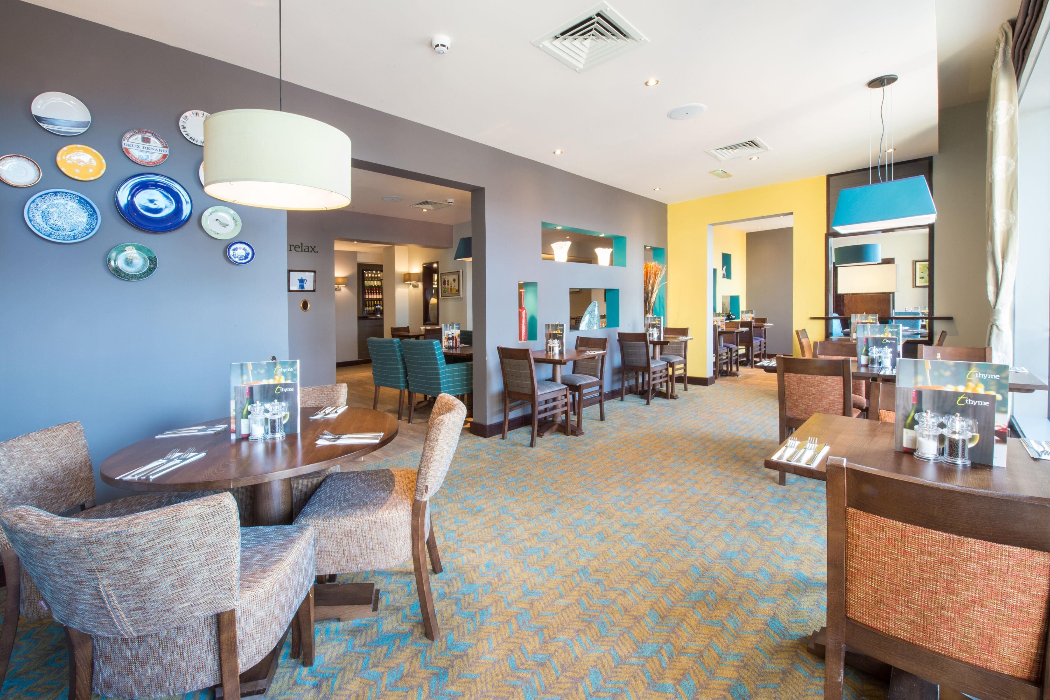 A hotel dining area with multiple tables and chairs, a patterned carpet, and wall-mounted plates.