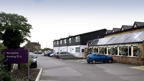 A parking area with a reception sign, a building with a sloped roof, and parked cars. - Premier Inn Cambridge North - Girton (Cambridge)