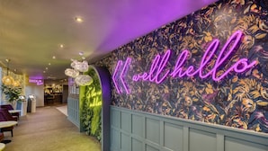 Una hall d'albergo con un'insegna luminosa che recita 'well hello', una reception e un banco informazioni.