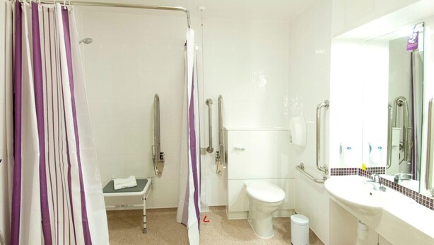 A bathroom with a shower, toilet, and sink, equipped with grab bars and a wheelchair-accessible bench. - Premier Inn Cwmbran (Cwmbran)