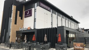 Egy épület, amelyen a „Premier Inn” és a „Beefeater” feliratok láthatók, és amelynek külső, napernyőkkel ellátott ülőhelye van.