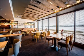 En restaurang med synliga träbjälkar i taket, stora fönster med persienner och utsikt över vattnet.