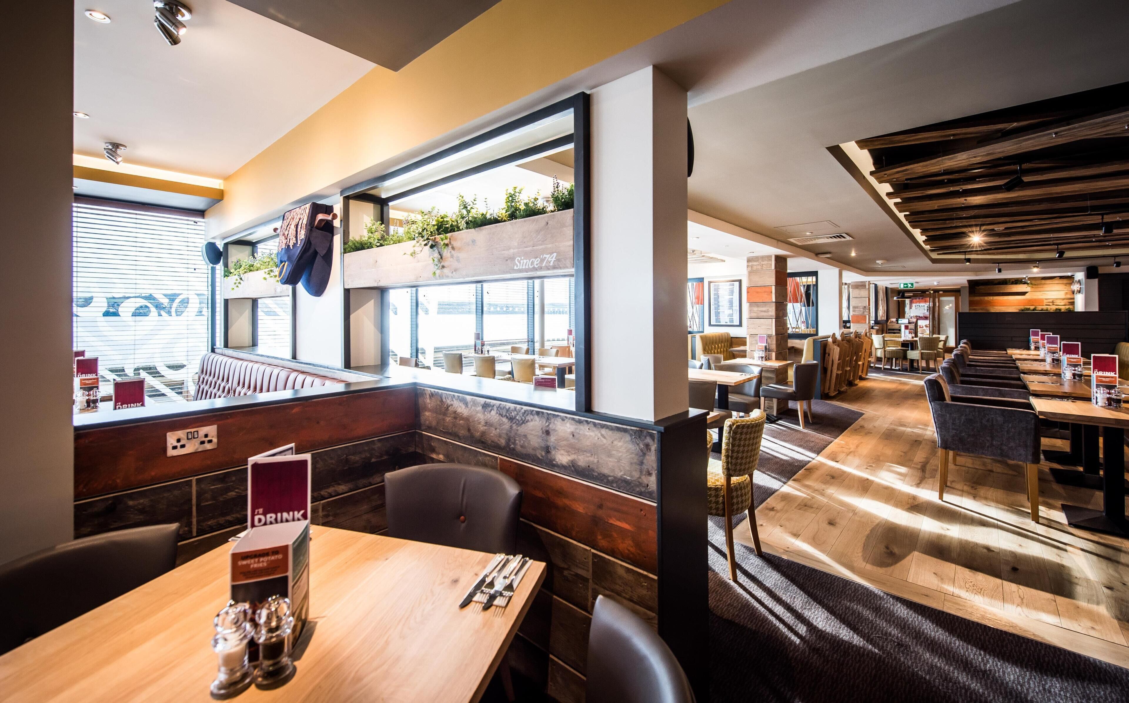 Un restaurante con un interior moderno, mesas de madera y vistas amplias a través de grandes ventanales.