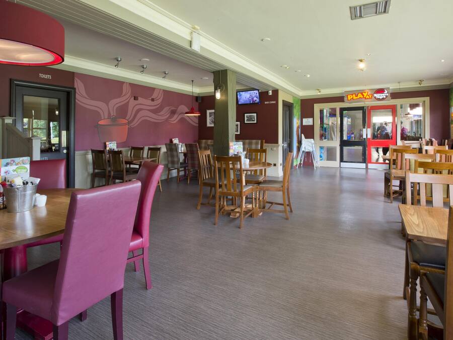 Interior de un restaurante con varias mesas y sillas, un televisor en la pared y un letrero que indica una zona de juegos.