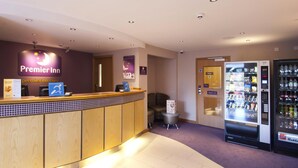 A hotel reception area with a wooden reception desk, a Premier Inn sign, a seating area, and vending machines. - Premier Inn Burton On Trent Central (Burton on Trent)