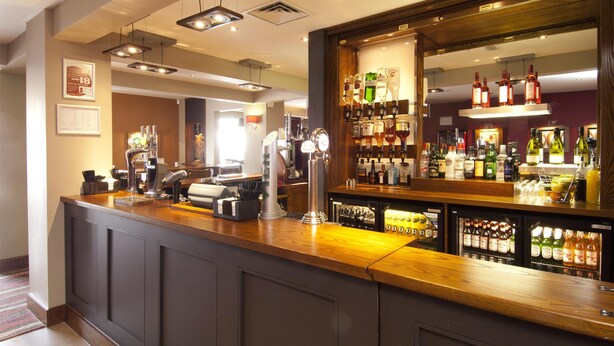 A bar area with a counter, bar stools, and a variety of bottles and glasses. - Premier Inn Exeter Central St Davids (Exeter)