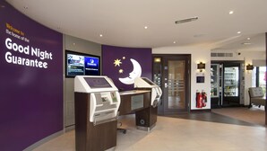 A hotel reception area with a purple wall, a digital screen, a check-in counter, and a glass door leading to the lobby. - Premier Inn Exeter - M5 J29 (Exeter)