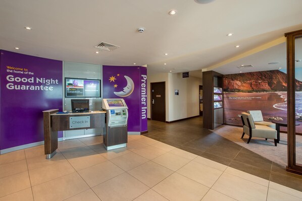 Un hall d’hôtel avec une pancarte indiquant une « garantie de bonne nuit », un comptoir d’enregistrement, le logo de Premier Inn et un espace détente agrémenté d’une fresque murale représentant un paysage.