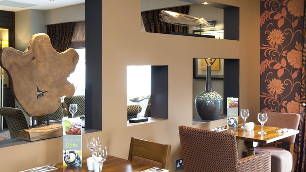 A dining area with a wooden table, patterned chairs, a large decorative piece on the wall, and a menu stand. - Premier Inn Camberley (Camberley)
