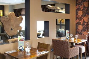 Un comedor con una mesa de madera, sillas estampadas, una pieza decorativa grande en la pared y un atril para el menú.