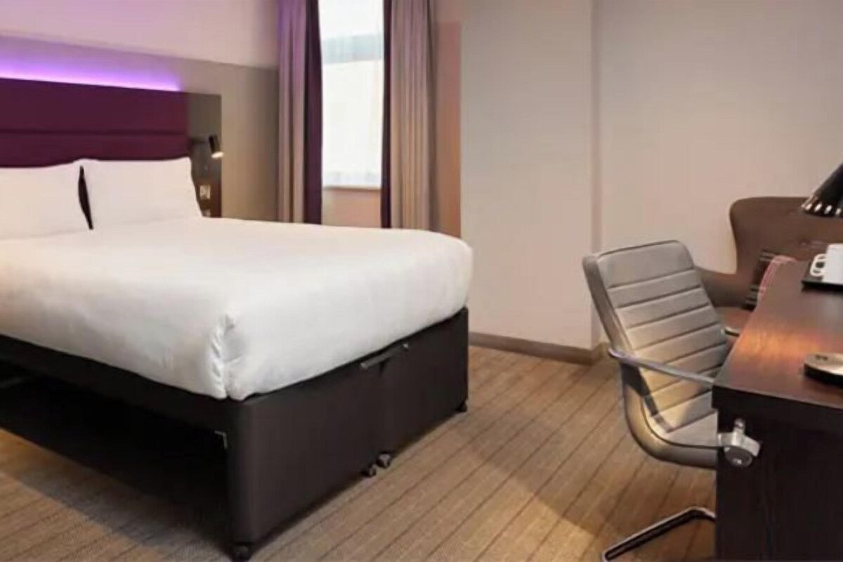 Quarto de hotel com cama, escrivaninha e cadeira, e janela com cortinas.