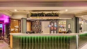 A reception area with a green counter, a wooden door, a wall with tropical wallpaper, and a bar area with a neon sign. - Premier Inn Glasgow Airport (Paisley)