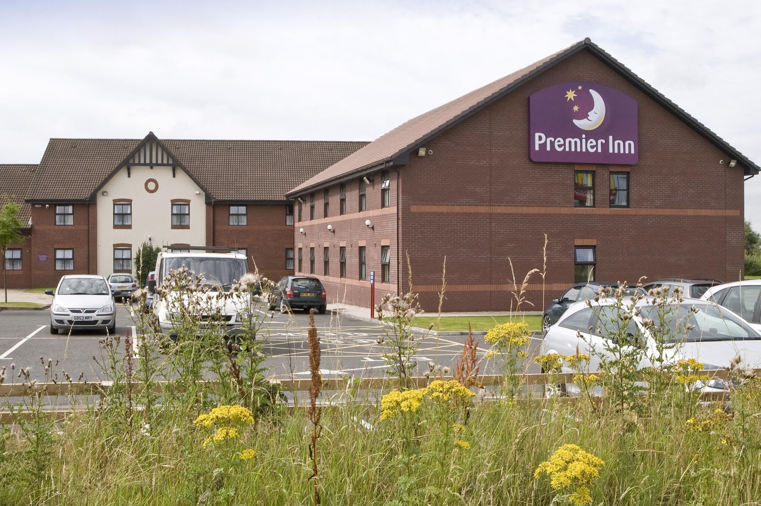 Et hotel med et tydeligt 'Premier Inn' skilt, en parkeringsplads med flere biler, og en bygning med facade i mursten.