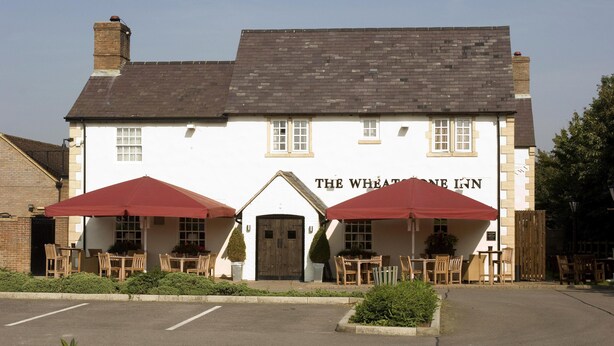 A white building with a sign reading 'The Wheatone Inn.' There are two red umbrellas providing shade over outdoor seating. - Premier Inn Gloucester - Barnwood (Gloucester)
