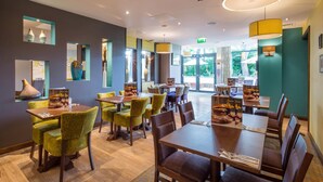 A restaurant with multiple tables and chairs, a ceiling with recessed lighting, and a large window providing a view of the outdoors. - Premier Inn Harrogate Town Centre (Harrogate)