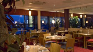 Un restaurant avec des tables et des chaises rondes, offrant une vue sur le paysage urbain grâce à de grandes fenêtres.