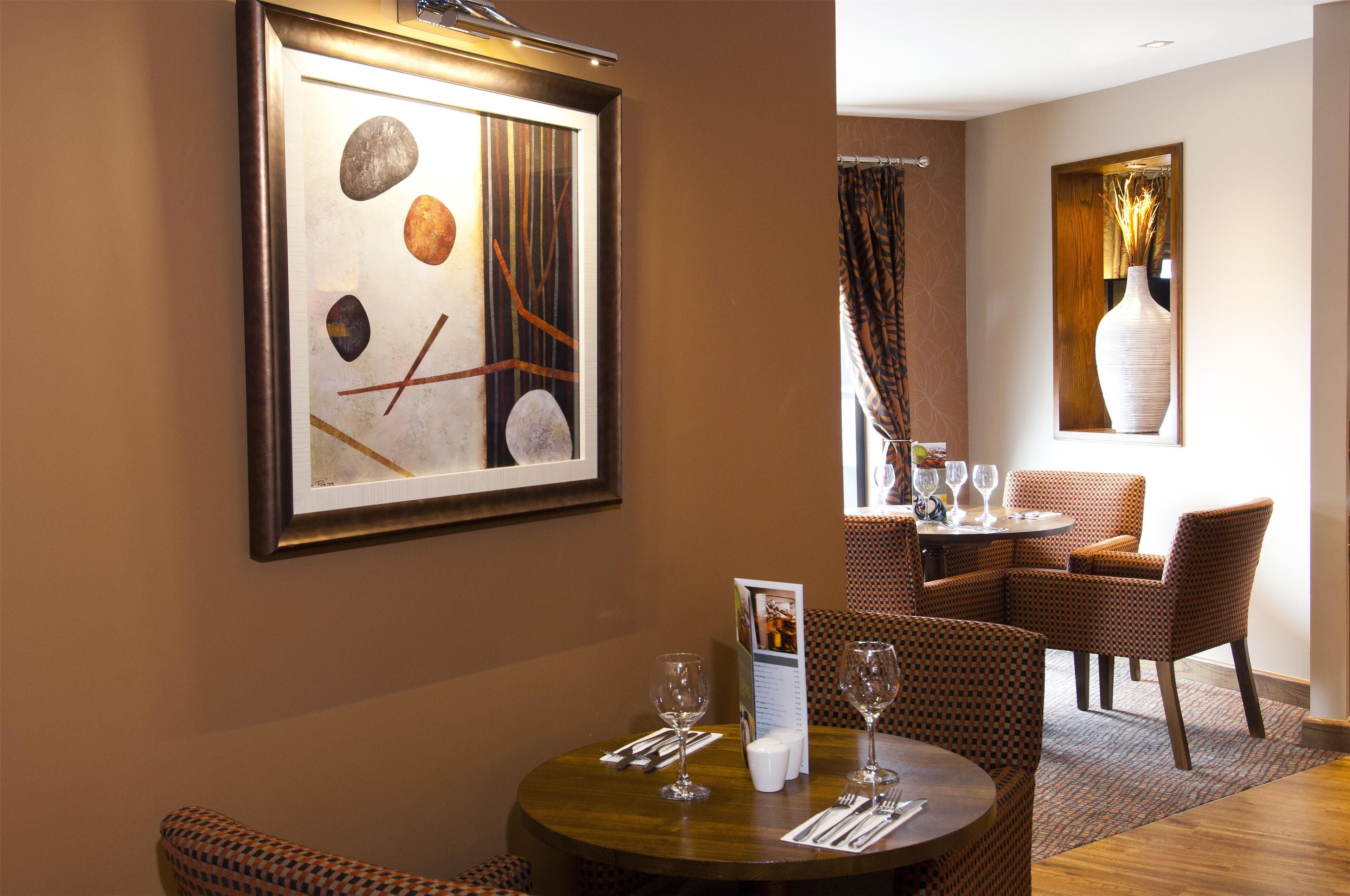A dining area with a round table, wicker chairs, a framed abstract painting, and a vase on the wall.