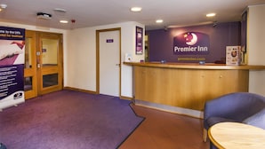 Un espace d’accueil d’hôtel avec un comptoir en bois, une enseigne « Premier Inn », une porte menant à une chambre privée, et un bureau de réception équipé d’un ordinateur et d’un tableau affichant le menu.