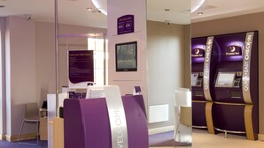 Ein Hotel-Empfangsbereich mit modernem Design, darunter eine Rezeption, ein digitales Check-in-Terminal und eine Anzeige mit Zimmerpreisen.