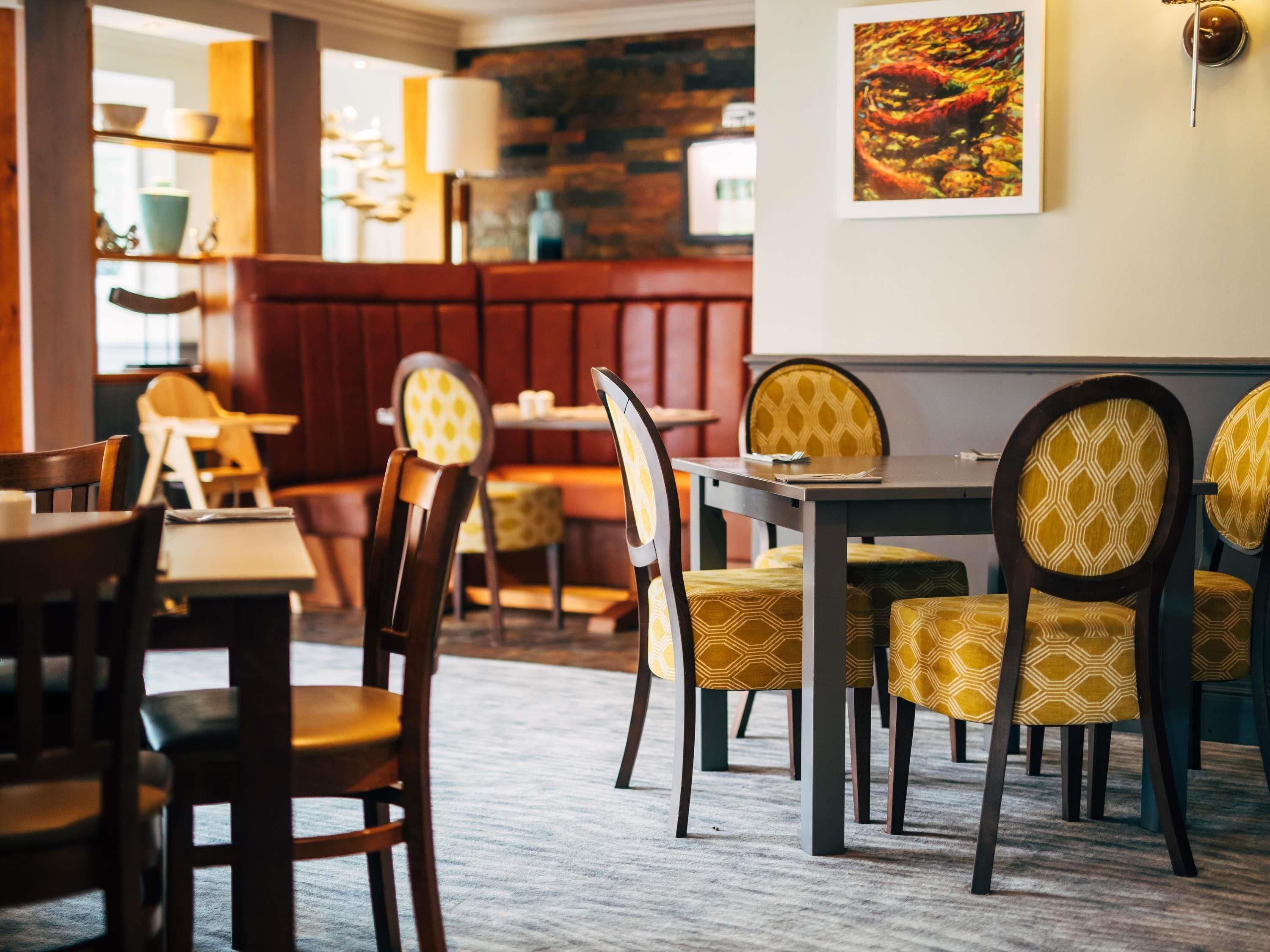 Interior de un restaurante con mesas y sillas de madera, cojines estampados y una pared de piedra.