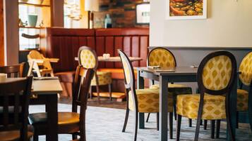 A restaurant interior with wooden tables and chairs, patterned cushions, and a stone wall.