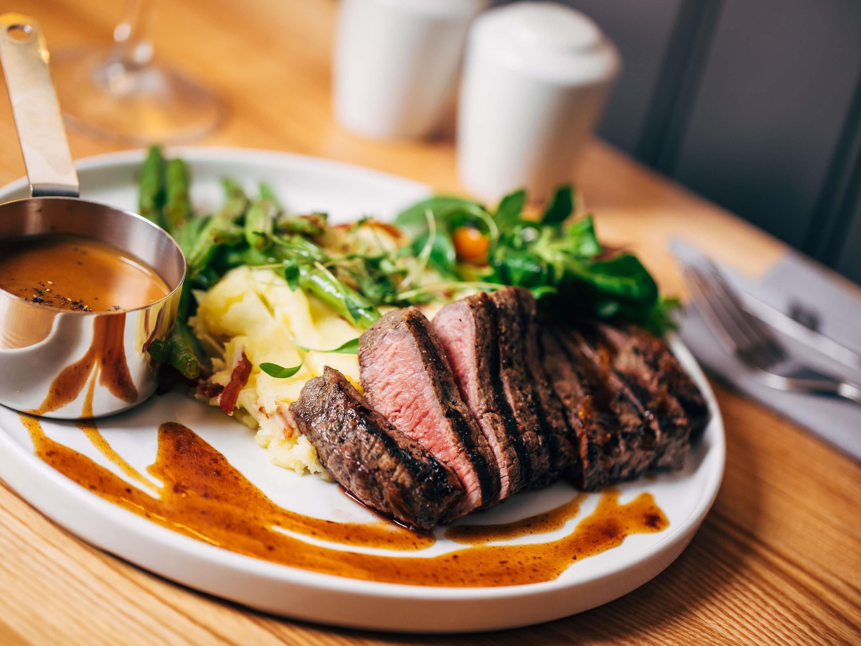Ein Teller mit Steak, Kartoffelpüree und Gemüse, eine Soßenkaraffe und ein Weinglas auf einem Holztisch.