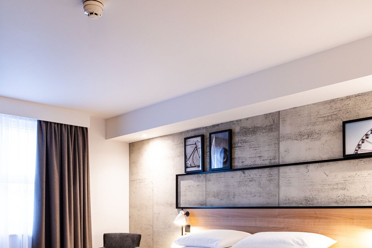 A modern hotel room with a large bed, a desk, and a chair. There are framed pictures on the wall and a window with curtains.