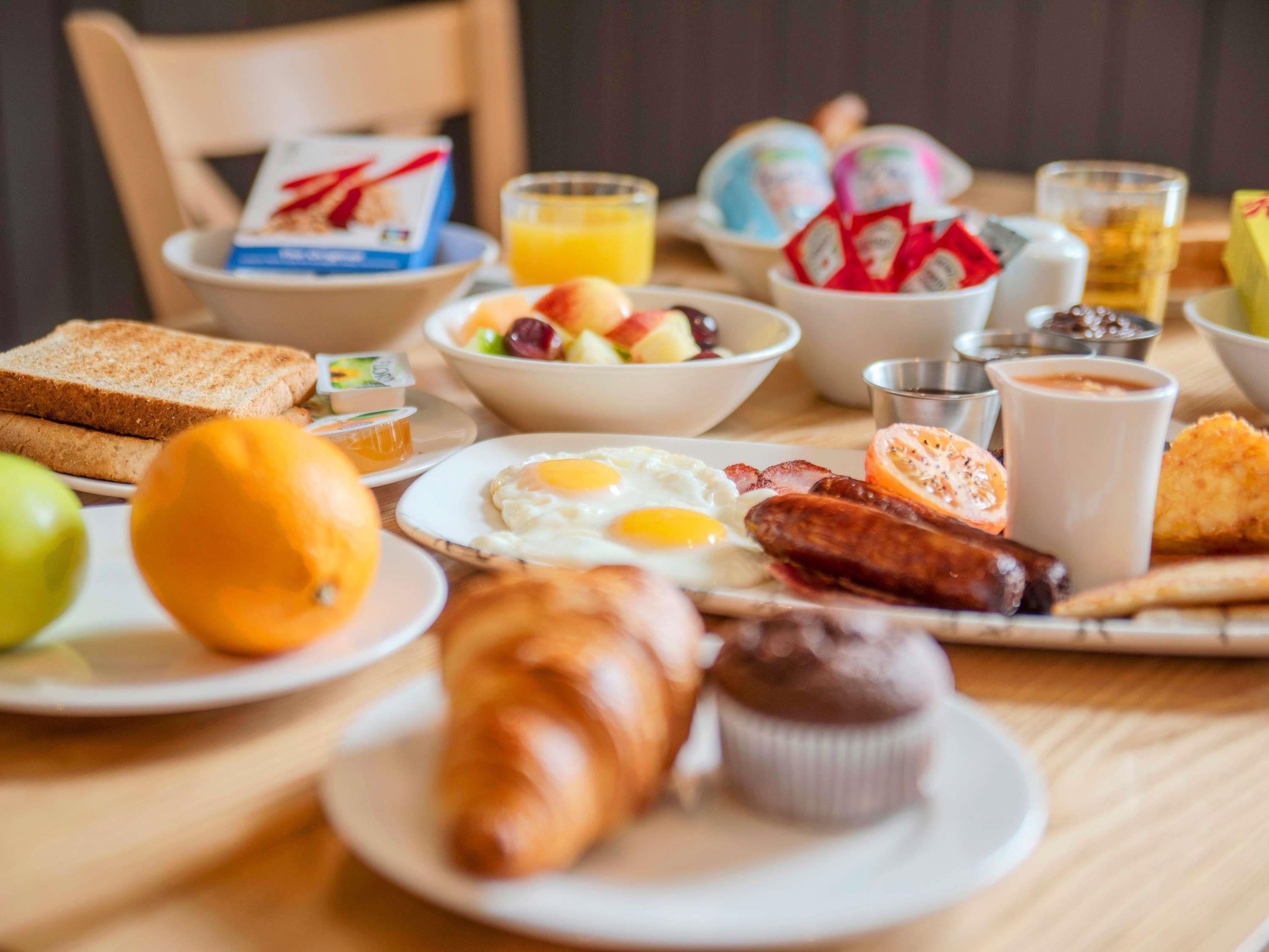 A breakfast table with toast, eggs, sausages, a croissant, a muffin, and a bowl of fruit.