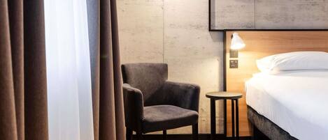 A hotel room with a bed, a chair, a small table, and artwork on the wall.