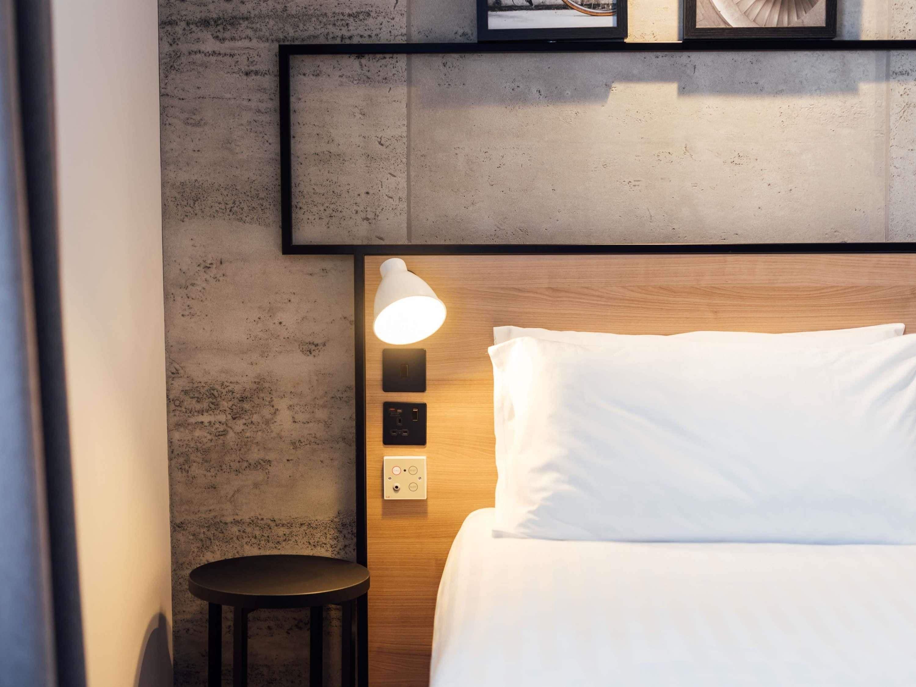 Una habitación de hotel con una cama, una mesita de noche con lámpara y un taburete pequeño.