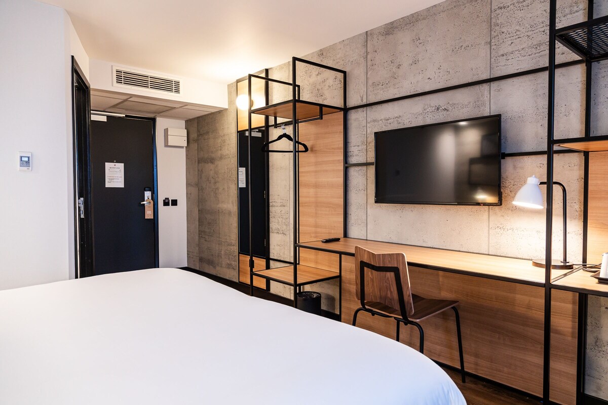 A modern hotel room with a large bed, a desk with a chair, a television, and a coat rack.