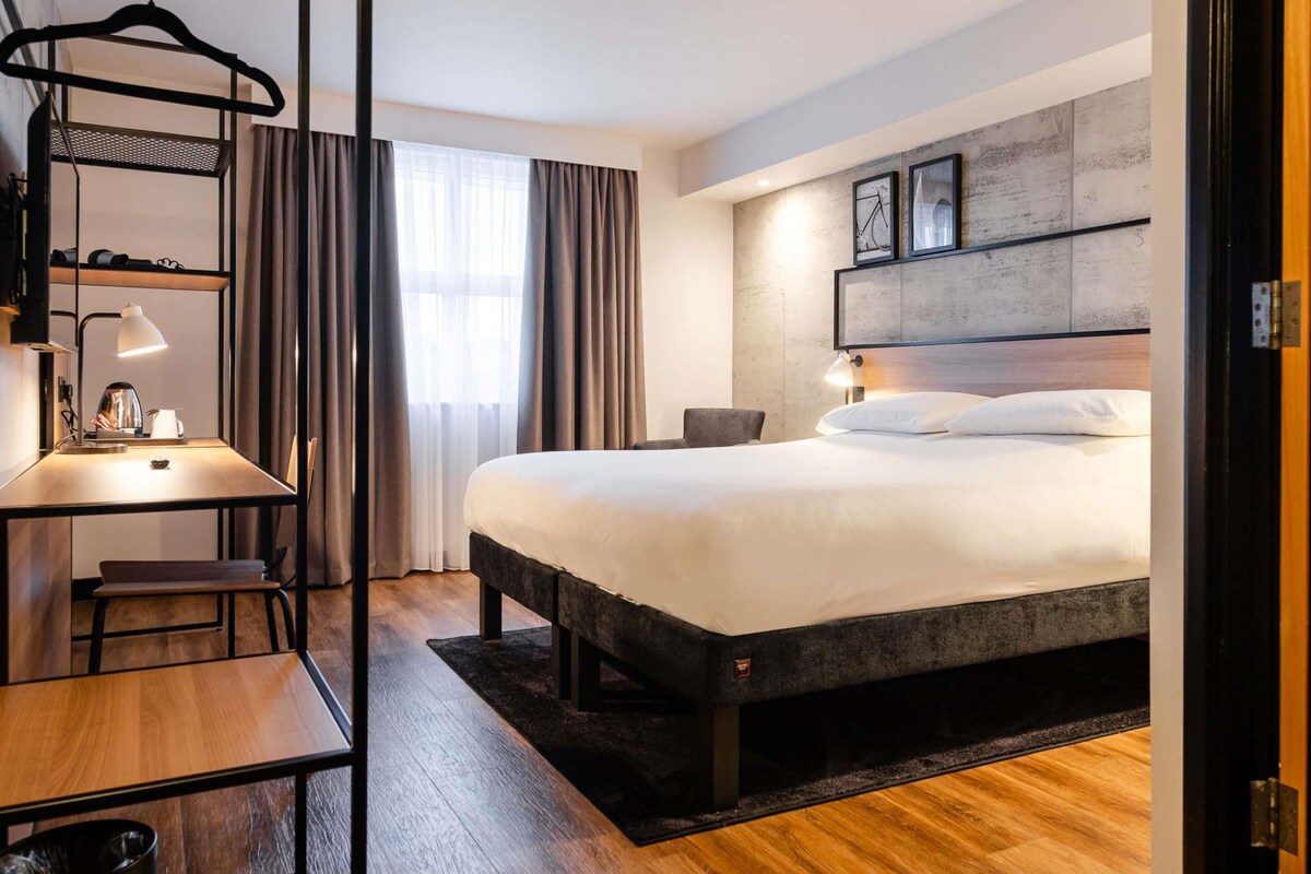 A modern hotel room with a bed, desk, and chair, a hanging rack, and a large window with curtains.