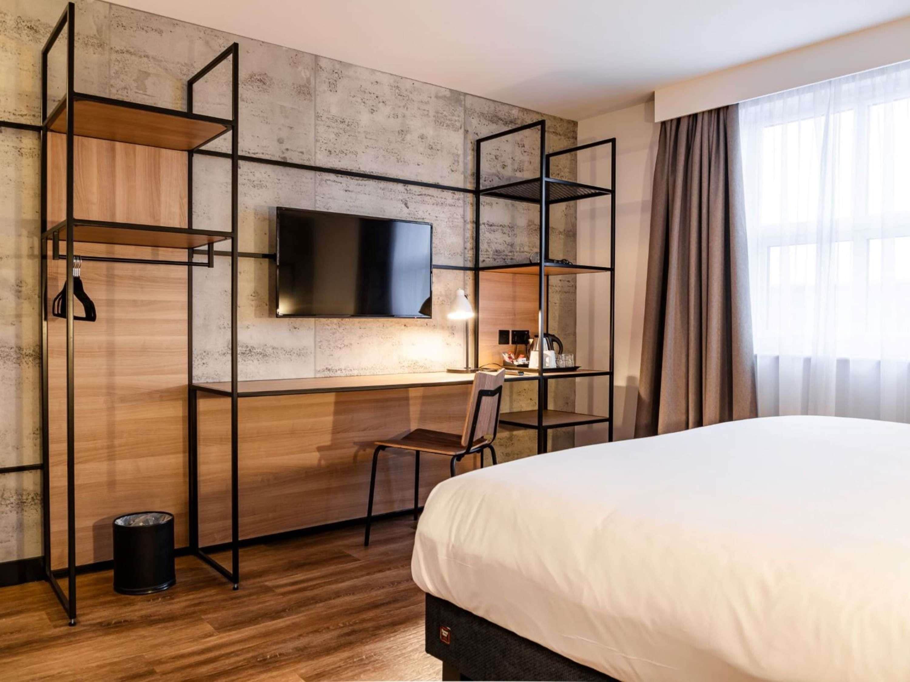 Camera d'albergo con un letto, una scrivania con una sedia, una TV montata su una parete strutturata e una finestra con tende.