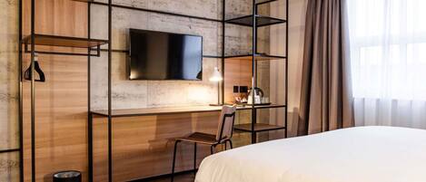 A hotel room with a bed, a desk with a chair, a TV mounted on a textured wall, and a window with curtains.