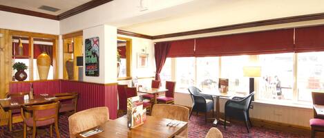 A restaurant with wooden tables and chairs, a patterned carpet, and large windows with red curtains.