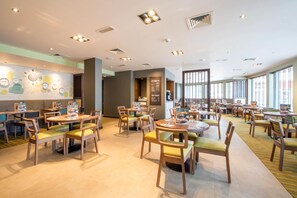 A spacious dining area with multiple tables and chairs, a wall with a clock and decorative elements, and large windows allowing natural light. - Premier Inn Leek Town Centre (Leek)