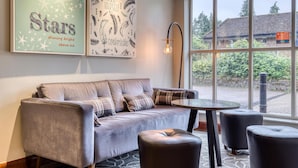 Ein modernes Wohnzimmer mit einem grauen Sofa, zwei schwarzen Hocker, einem runden Tisch und einem großen Fenster mit Blick auf Grünflächen und ein Gebäude.