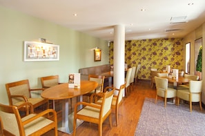 A dining area with wooden tables and chairs, a patterned wallpaper wall, and a large window. - Premier Inn London Gatwick Manor Royal (Crawley)