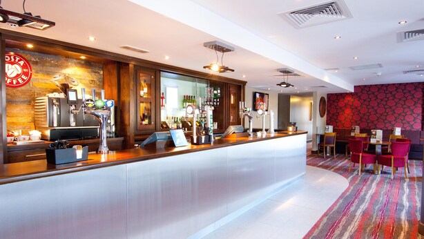 Een bar met een toonbank, meerdere tappunten en een ruim assortiment flessen. Er is een zithoek met tafels en stoelen.