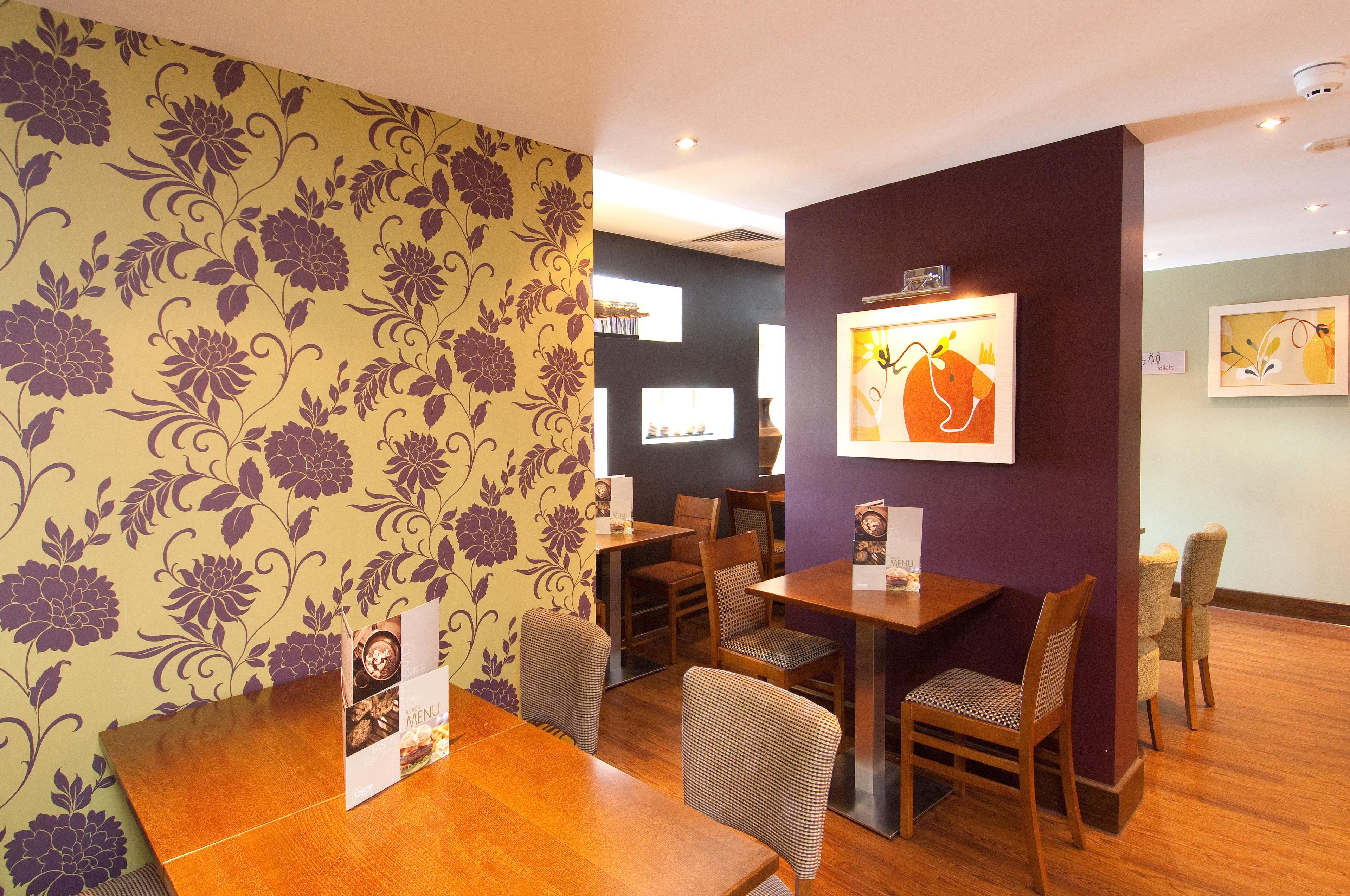 Un interior de restaurante con mesas y sillas de madera, papel tapiz floral y obras de arte enmarcadas en las paredes.