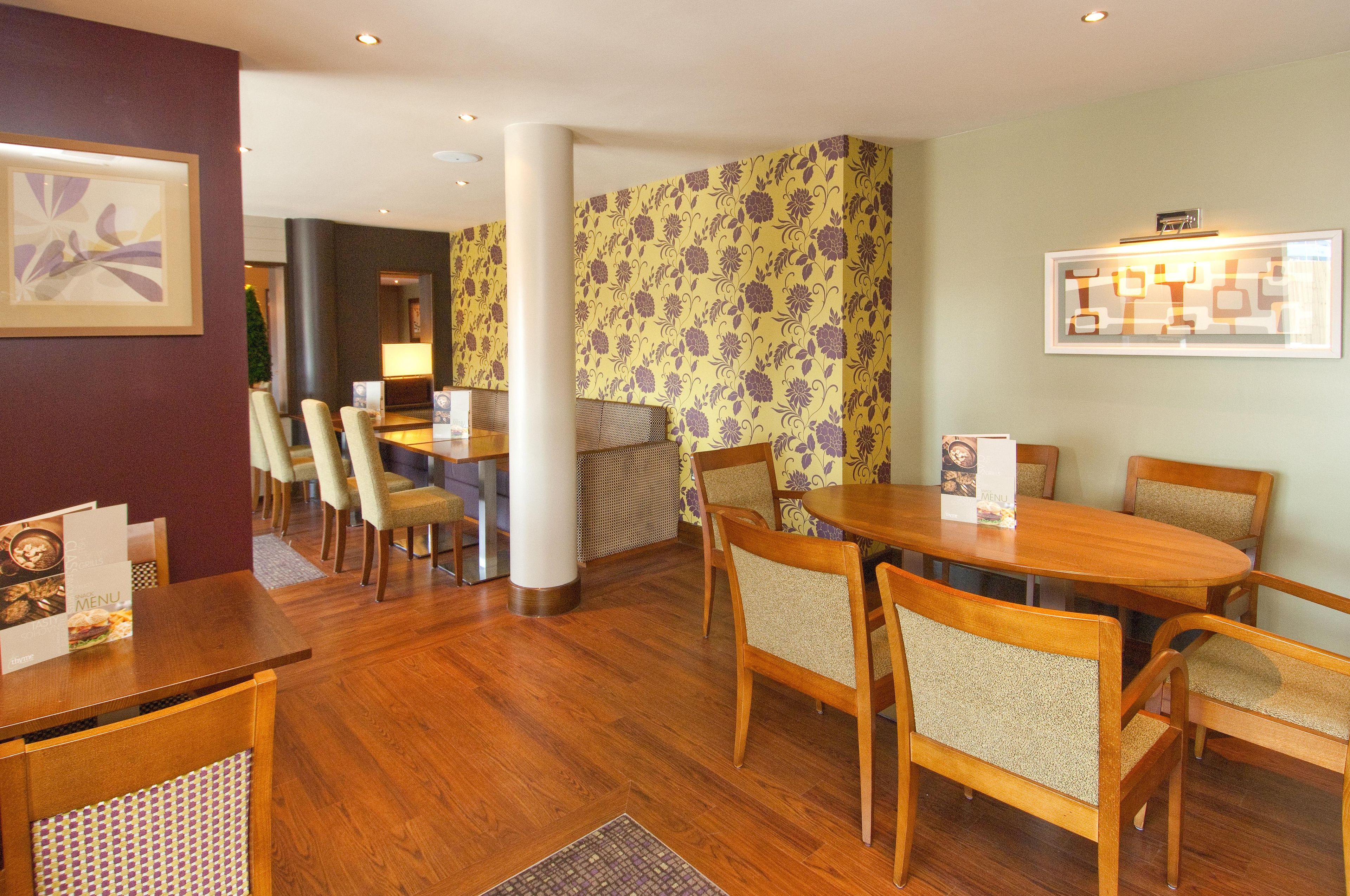 A hotel dining area with wooden tables and chairs, a reception desk, and a wall with a floral pattern.