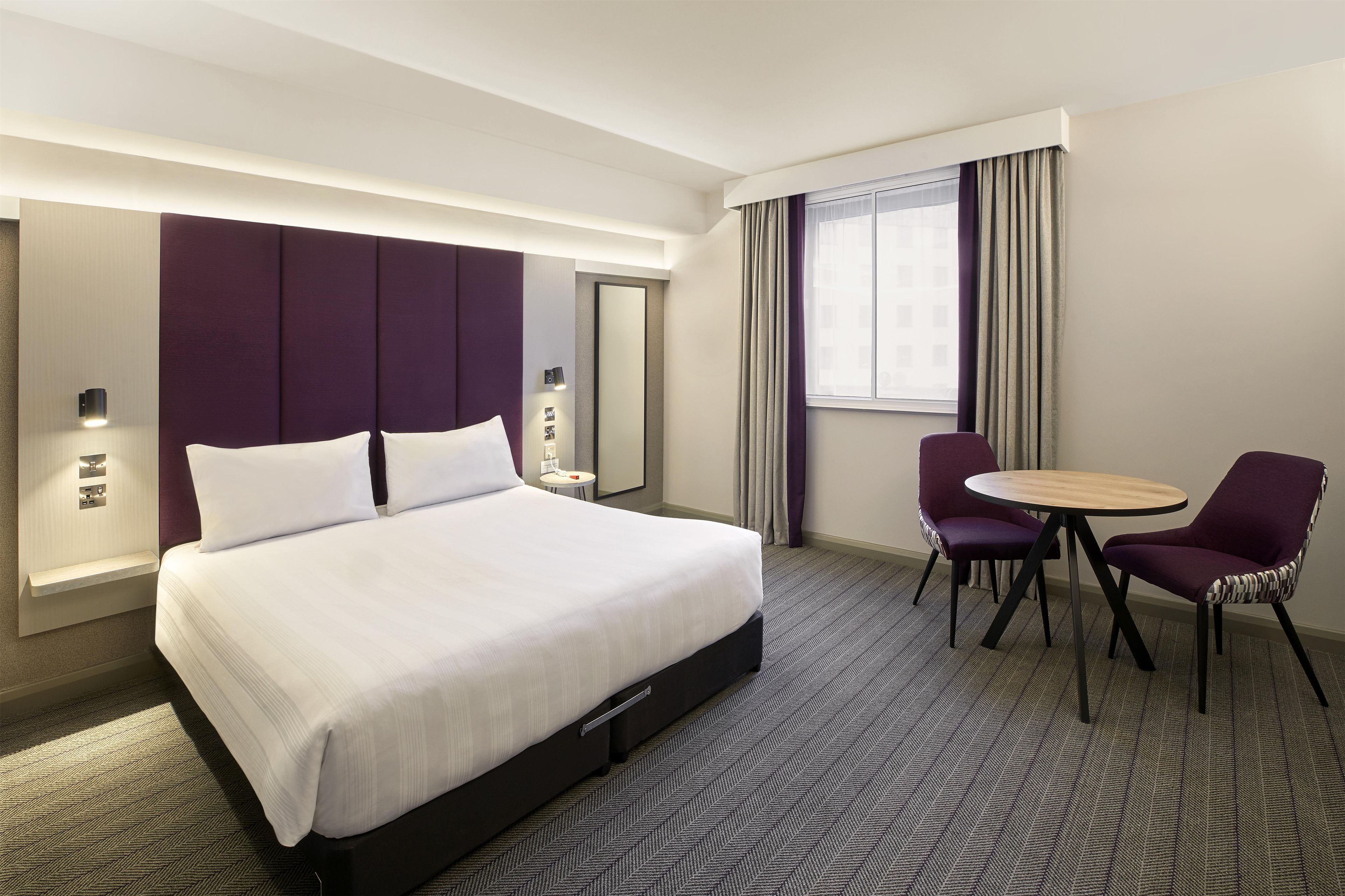 A hotel room with a large bed, a round table, two chairs, a mirror, and a window with curtains.