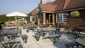 A patio area with multiple wooden tables and benches, a brick building with a tiled roof, and a hanging plant. - Premier Inn Colchester - A12 (Colchester)