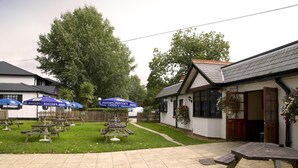 A courtyard with picnic tables and umbrellas, a white building with a wooden door, and a large tree. - Premier Inn Cobham (Cobham)